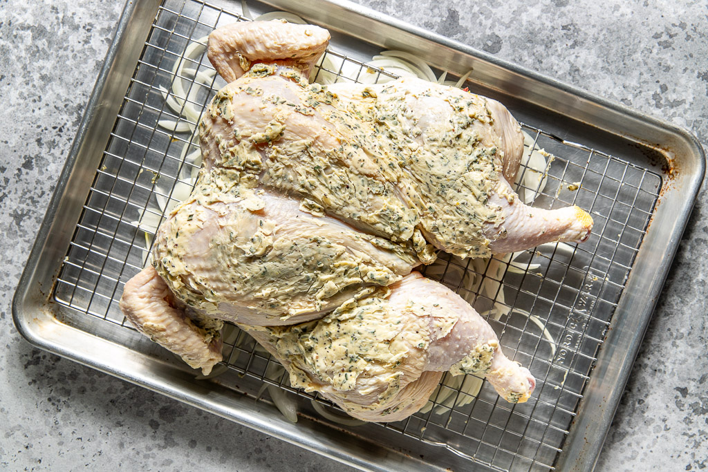 Raw spatchcocked chicken coated in herb butter on a wire rack over a baking sheet with sliced onions on it.