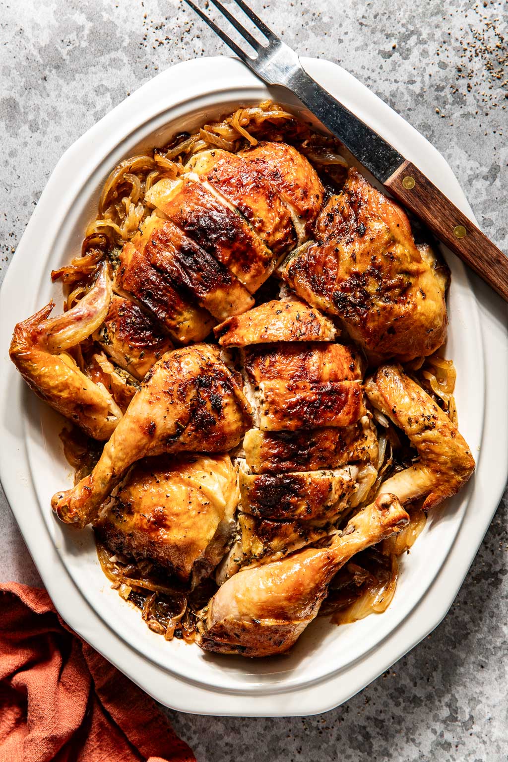Roast chicken cut into pieces on a white platter with caramelized onions, next to utensils and drinks.