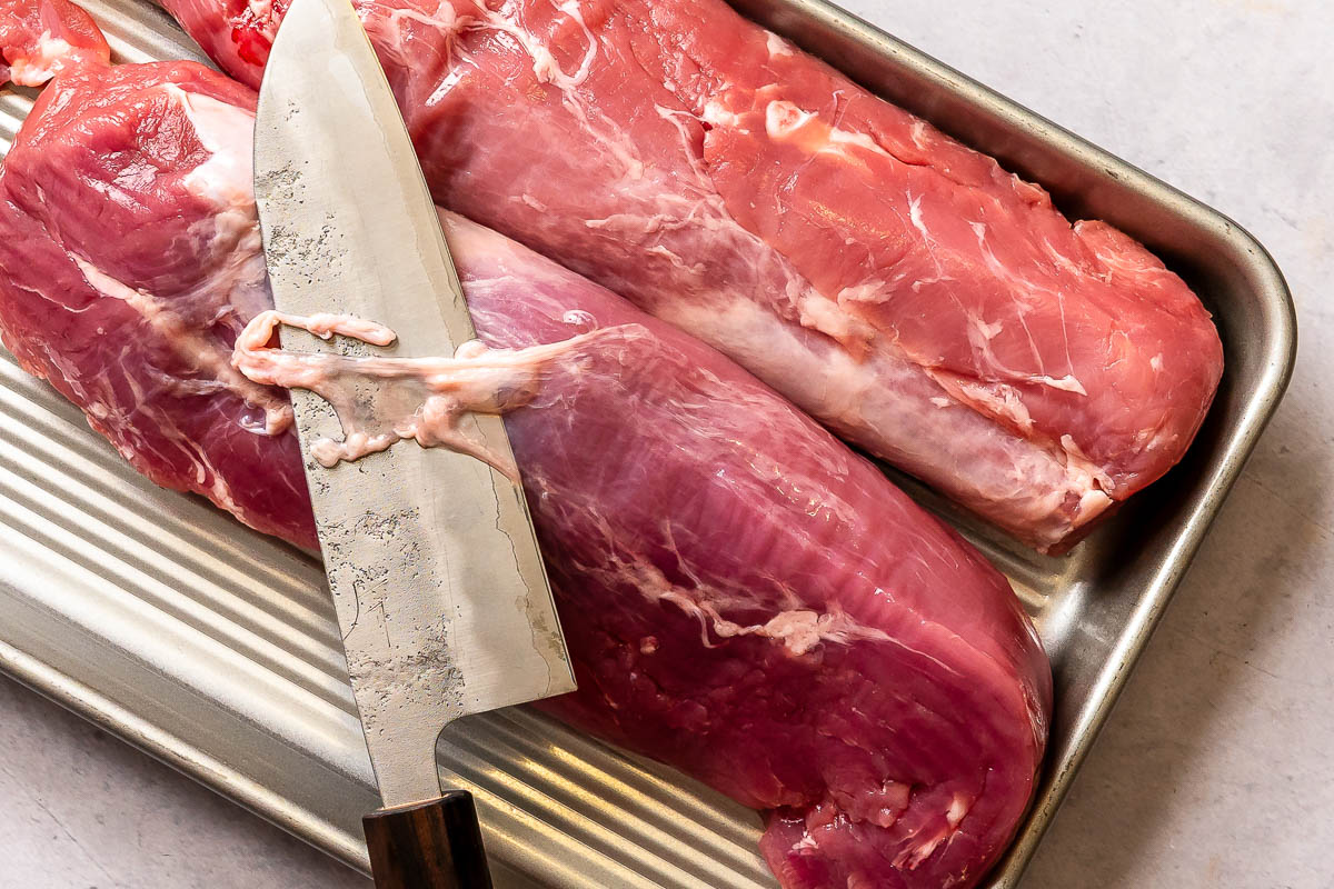 Pork tenderloin being trimmed with a large chefs knife.