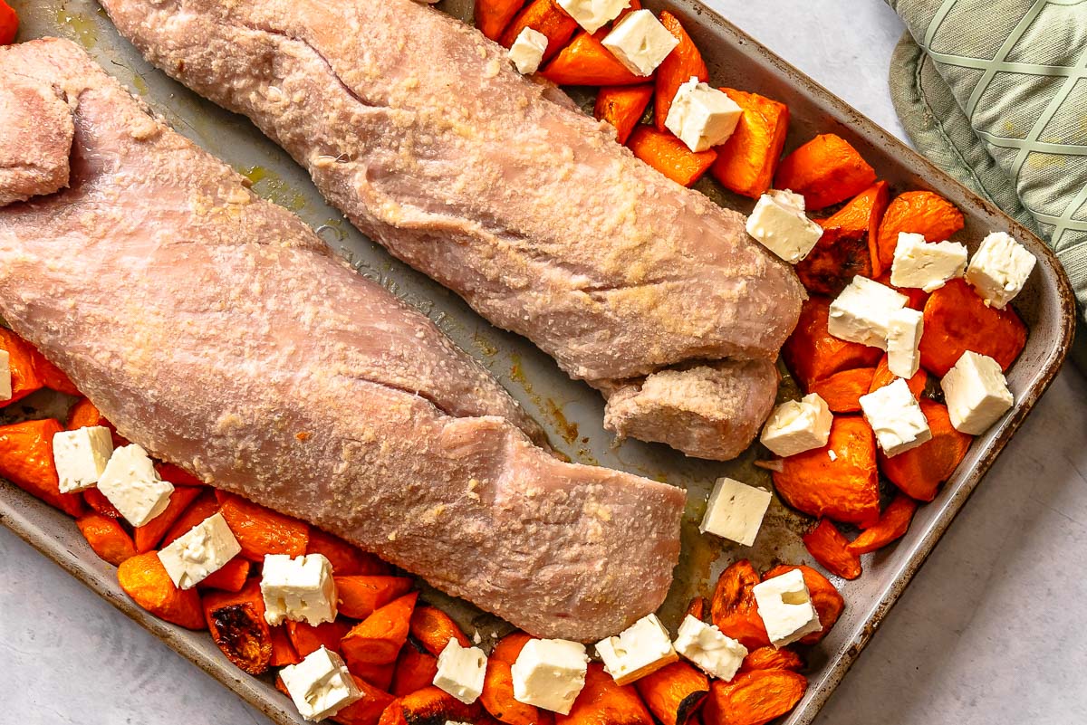 Partially-cooked pork tenderloin on a baking sheet with cubed carrots and chunks of feta.