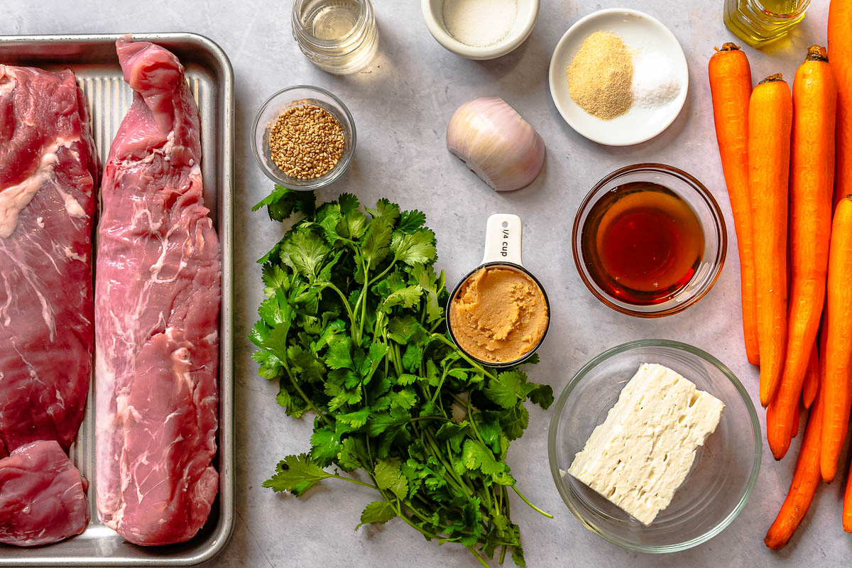 Raw pork, cilantro, miso, sesame seeds, vinegar, onion, garlic powder, salt, feta, toasted sesame oil and carrots set out on a counter.