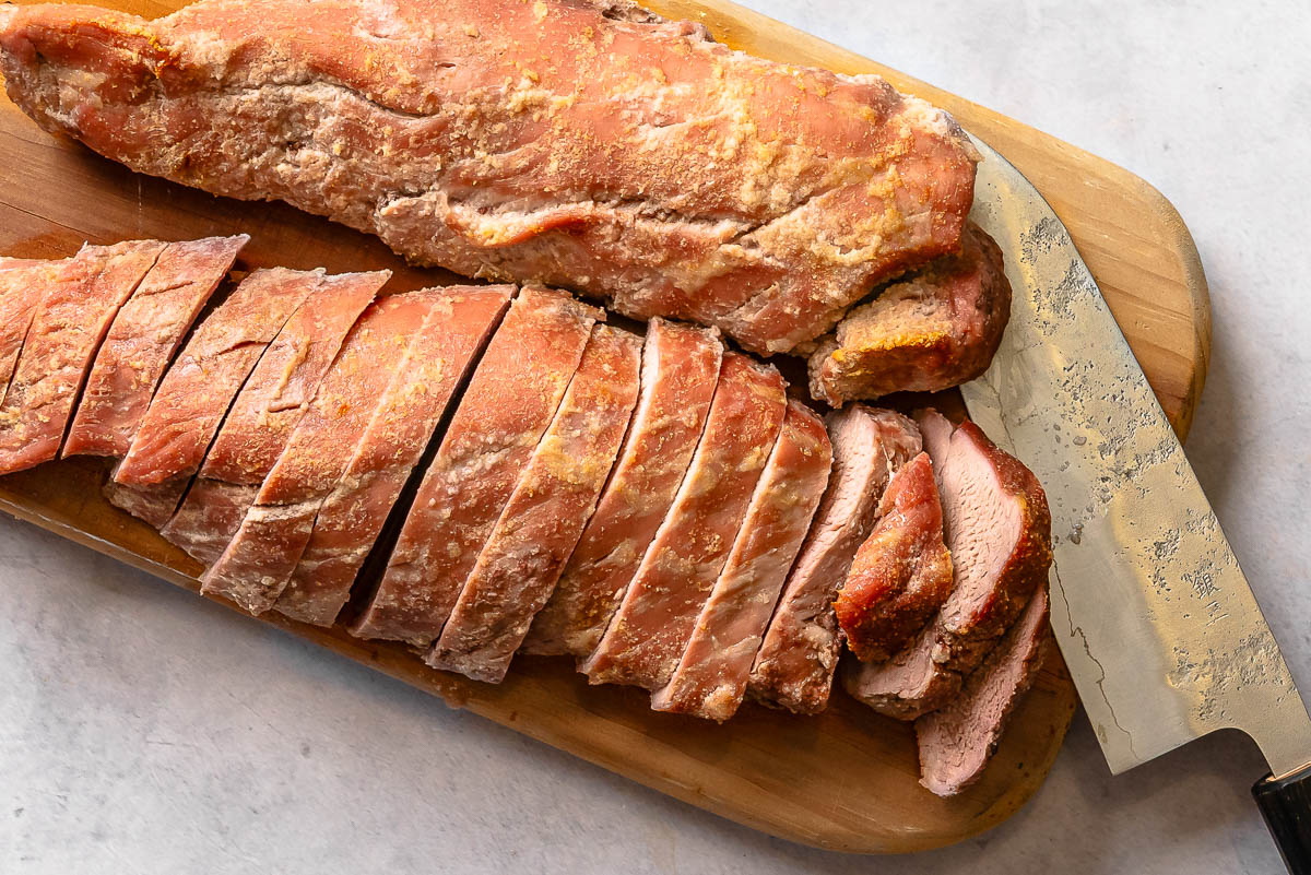 Sliced cooked pork tenderloin on a wooden cutting board next to a large knife.