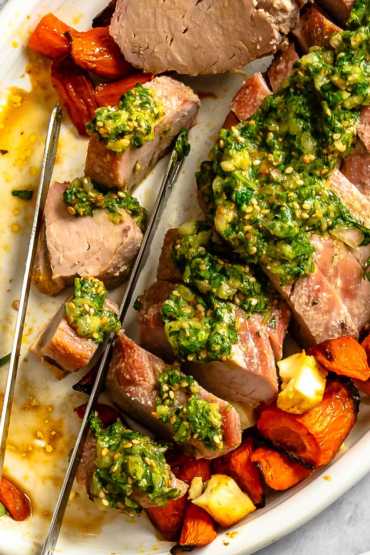 Sliced pork tenderloin topped with green herb sauce, served with roasted carrots and feta on a white plate.