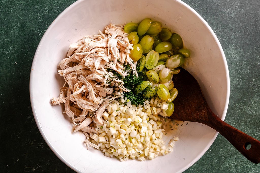 Shredded chicken, halved green grapes, chopped celery, and herbs in a white bowl with a dark wooden spoon set in it.