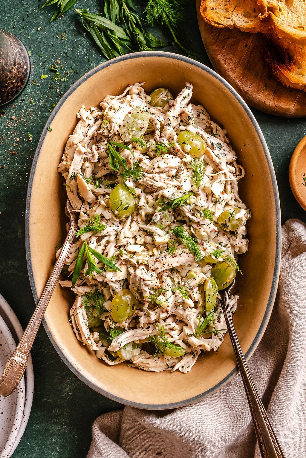 Oval bowl of creamy chicken salad with green grapes and fresh herbs, served with toasted bread on the side.