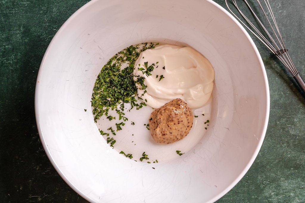 Mayonnaise, mustard and tarragon in a large white mixing bowl with a whisk set nearby.