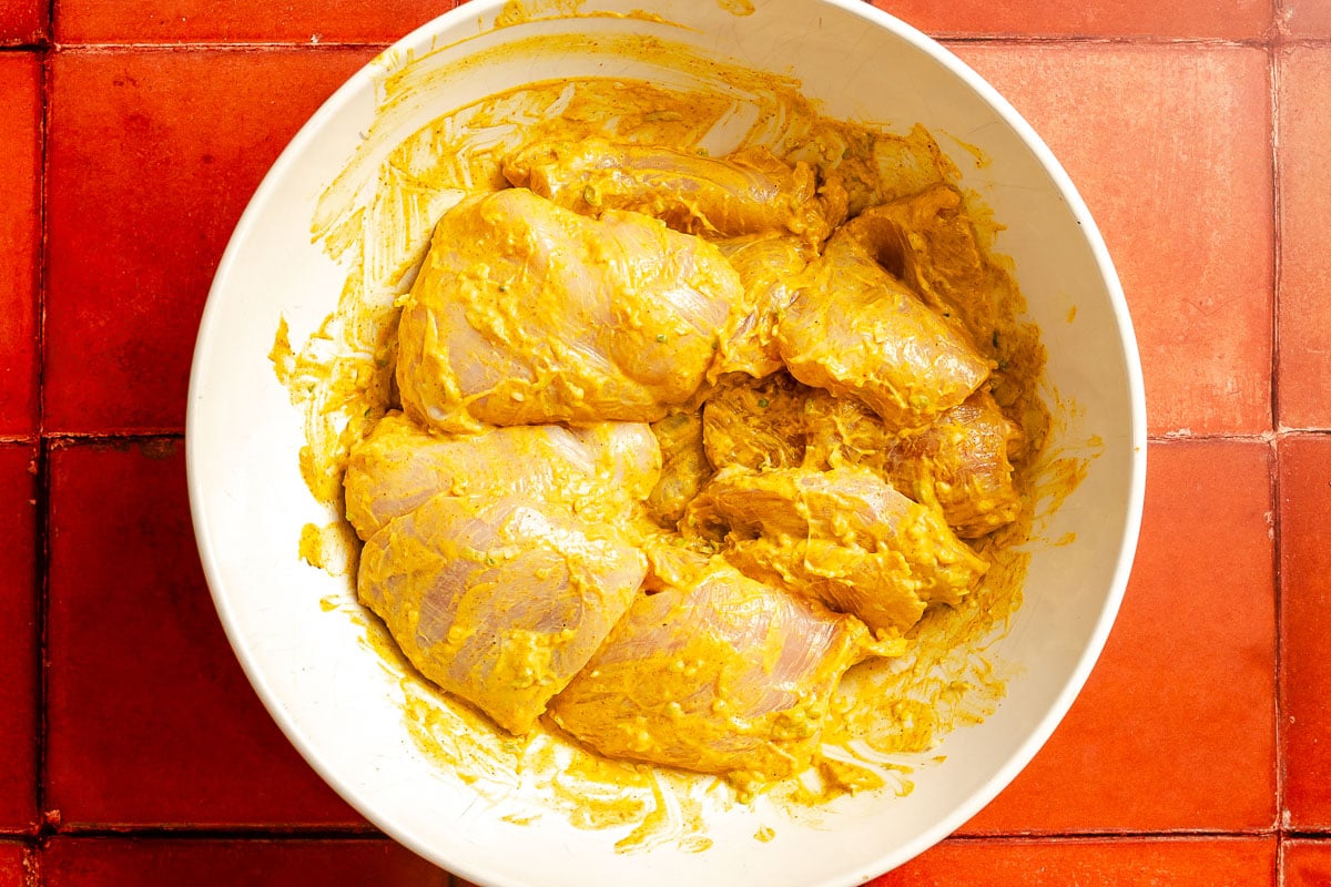 Raw chicken breast pieces marinate in a creamy curry marinade in a large white mixing bowl.
