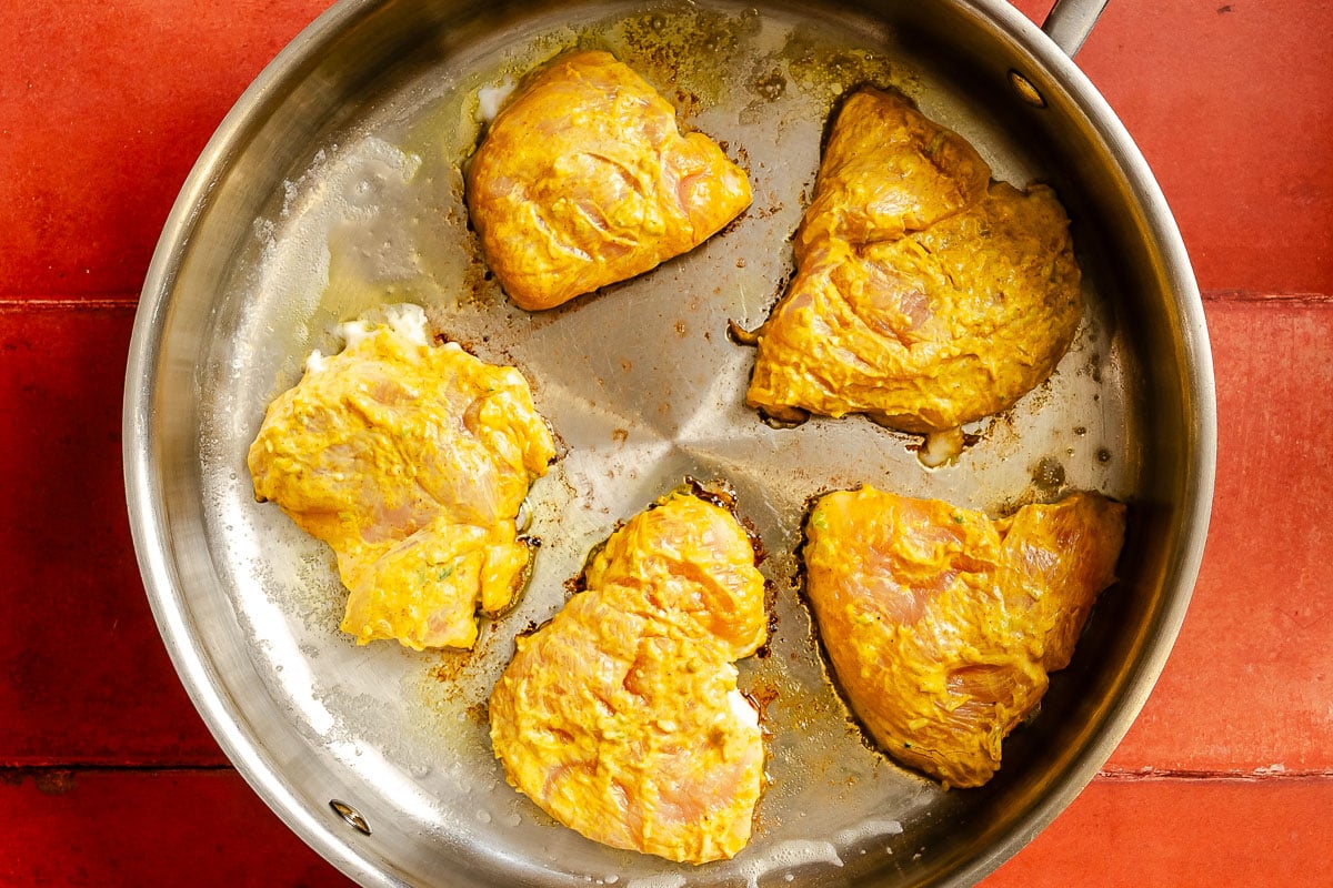 Five chicken breast pieces, marinated in curry, sizzle in a stainless steel skillet on a red tiled surface.
