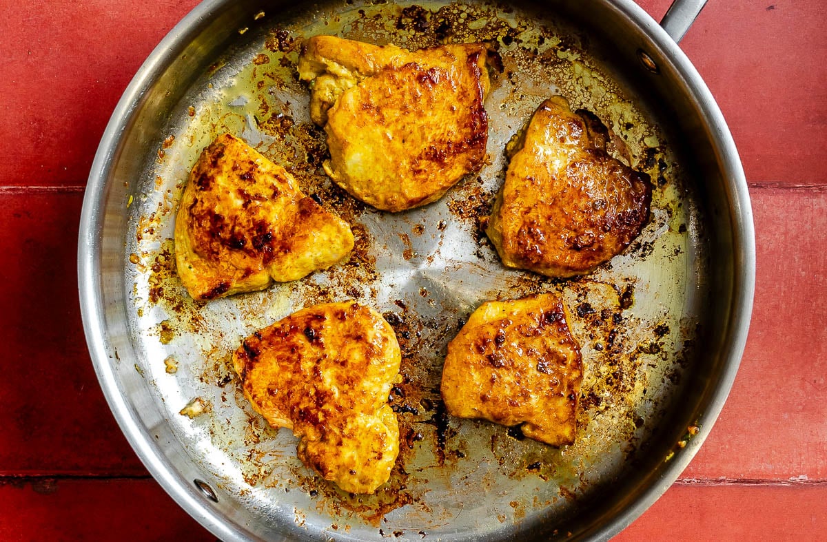 Five browned chicken breast pieces, marinated in curry, sizzle in a stainless steel skillet on a red tiled surface.