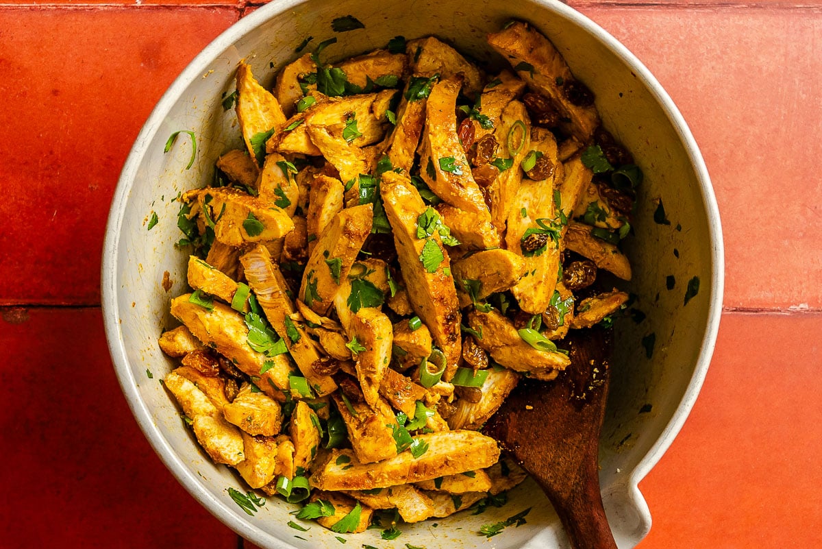Sliced curry marinated chicken breast tossed with cilantro and raisins in a large white mixing bowl.