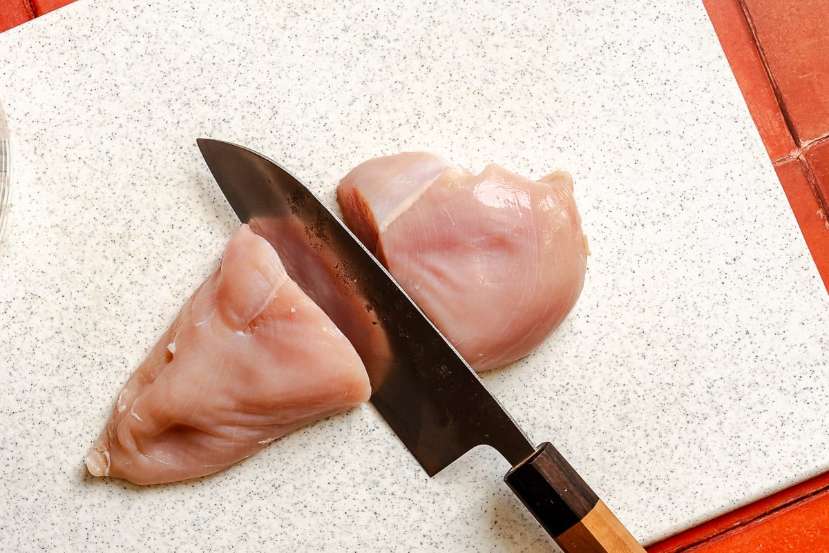 Raw chicken breast getting cut in half on a plastic cutting board with a chefs knife.