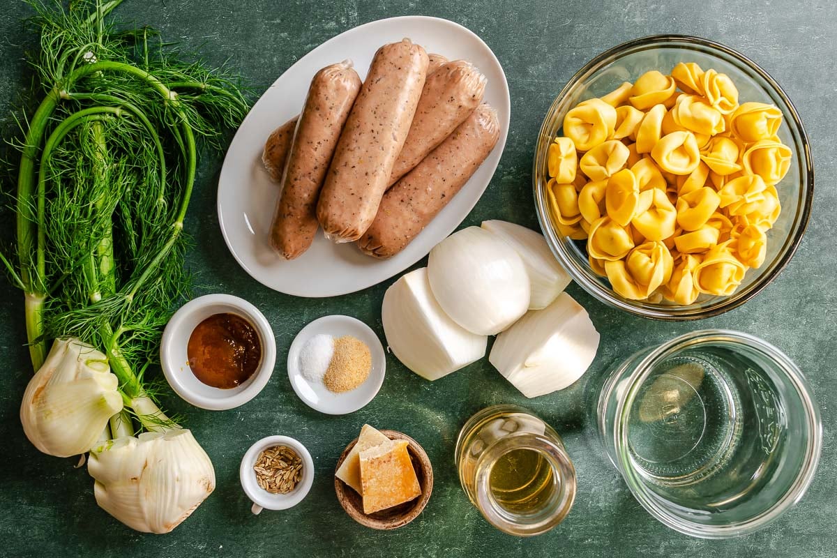 Fresh fennel, sausage, onion wedge, tortellini, Parmesan rinds, fennel seeds, granulated garlic, better than bouillon, wine, and water set out on a green counter.