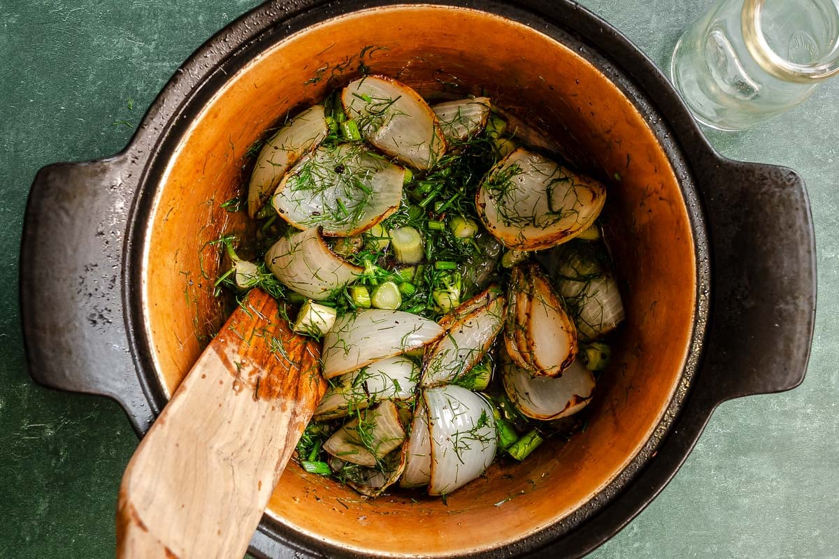 Onion wedges, chopped fennel stalks, and fennel fronds being sautéed in a large pot with a wooden spatula on the side.