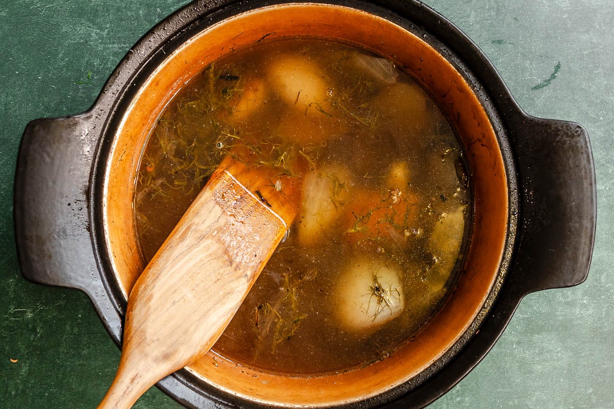 A pot of broth with fennel fronds, chopped fennel stalks, onion wedges, and a wooden spatula on a green surface.