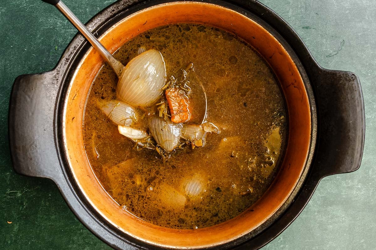 A pot of broth with fennel fronds, chopped fennel stalks, onion wedges, and a wooden spatula on a green surface.