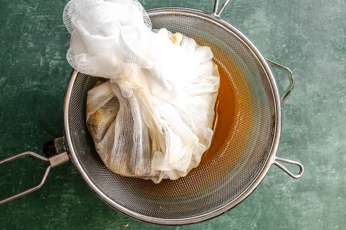 A bundle of cheesecloth in a strainer over a bowl, draining golden liquid on a green surface.