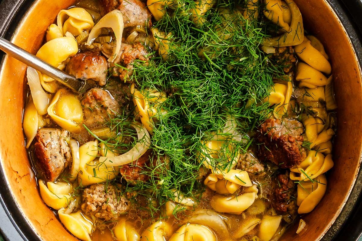 Chopped fennel fronds over top a pot filled with brothy tortellini, sausage and sliced fennel.