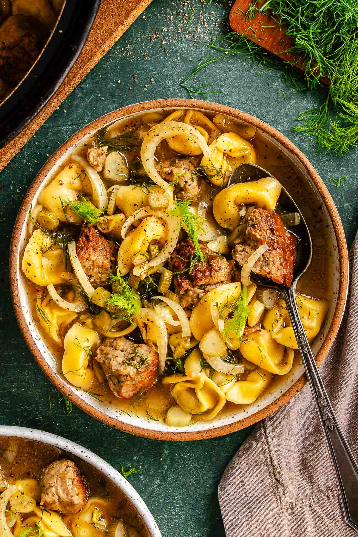 Bowls of brothy tortellini, sausage, and fennel, set on a green counter with fennel fronds scattered over top.