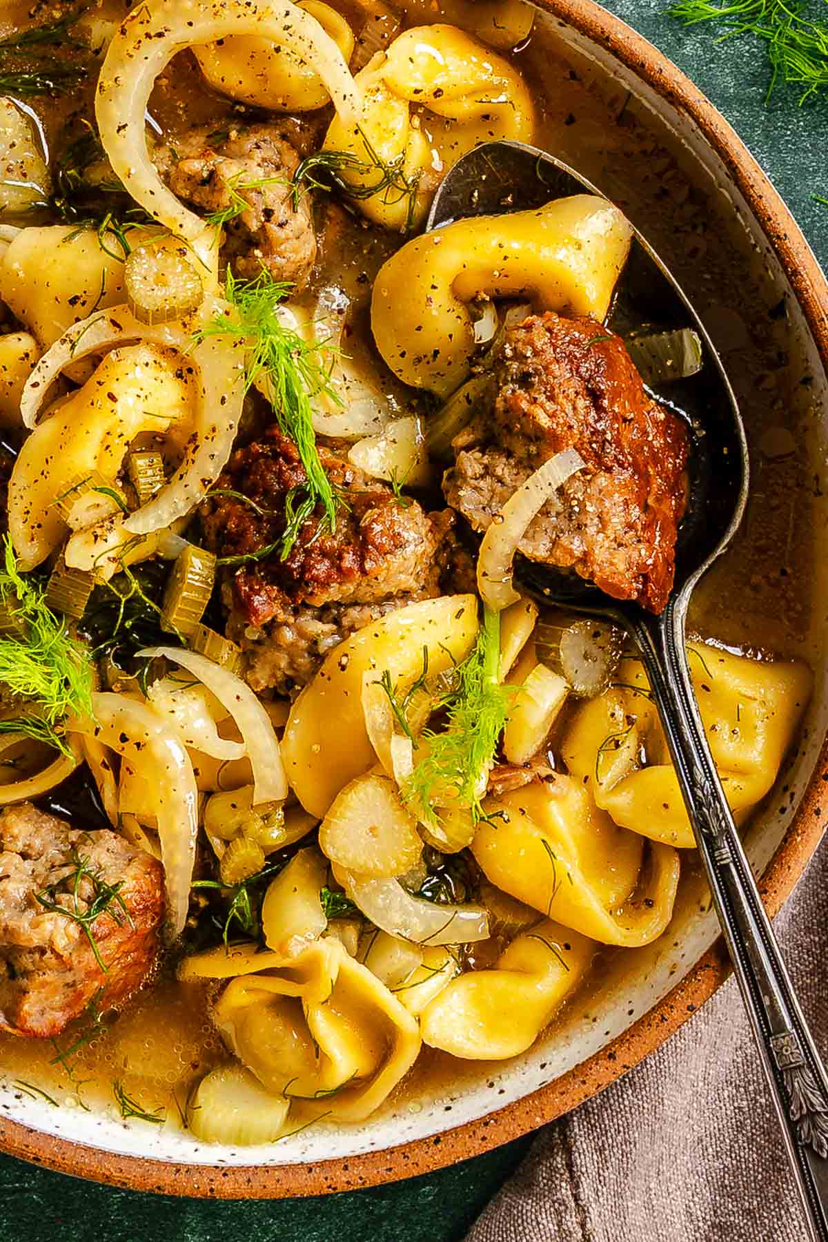 Bowls of brothy tortellini, sausage, and fennel, set on a green counter with fennel fronds scattered over top.