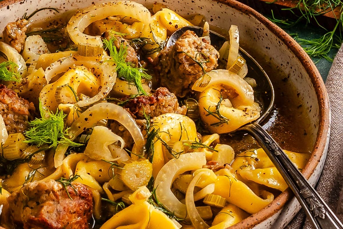Bowls of brothy tortellini, sausage, and fennel, set on a green counter with fennel fronds scattered over top.
