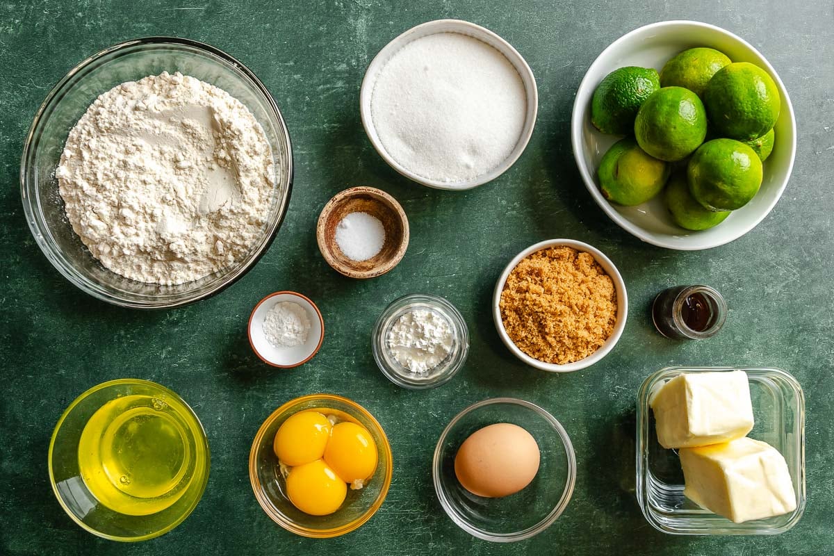 Flour, sugar, limes, salt, cornstarch, cream of tartar, brown sugar, vanilla, egg whites, egg yolks, an egg, and butter all set out on a green surface.
