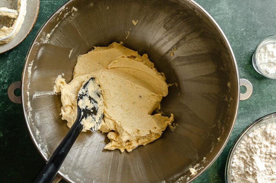 A mixing bowl with cookie dough set on a counter with a spoon set in th bowl. Mixer paddle attachment, flour and cornstarch set around.