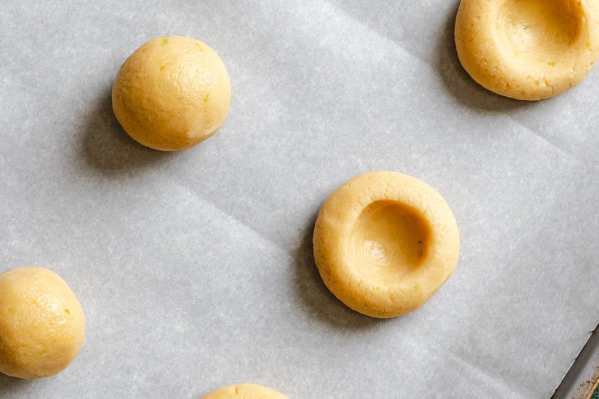 Four unbaked cookie dough balls, one with a thumbprint, on a sheet of parchment paper.