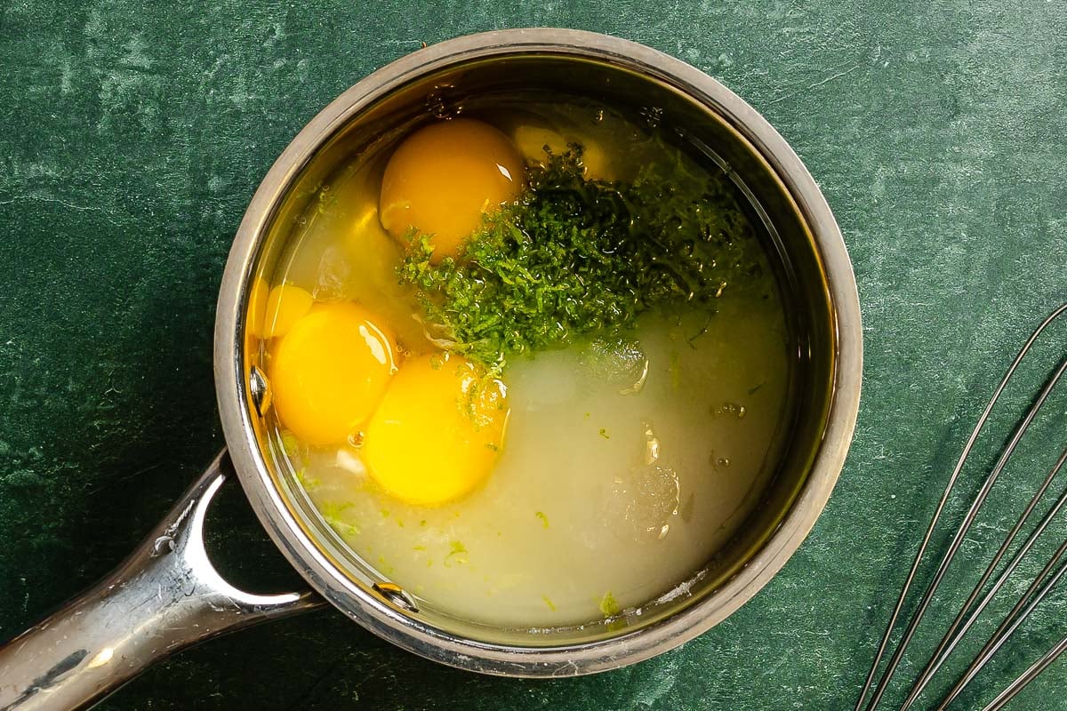 A saucepan with eggs, egg yolk, sugar, grated zest, and lime juice mixture set on a green surface.