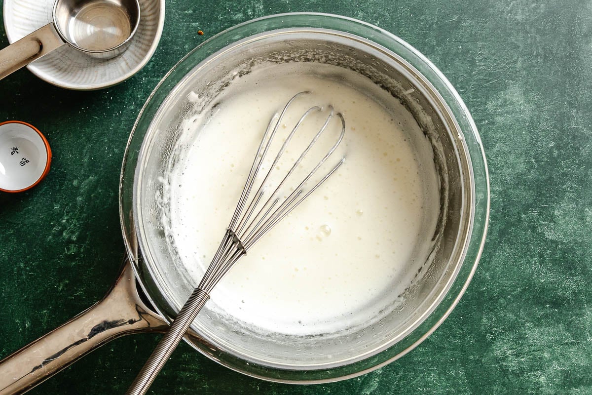 Cooked egg white and sugar meringue in a glass bowl with a whisk set over a saucepan.