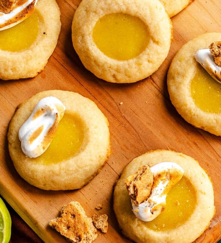 Round thumbprint cookie filled with yellow lime curd, some topped with meringue and graham cracker, sitting on a wooden board with crumbs scattered around.