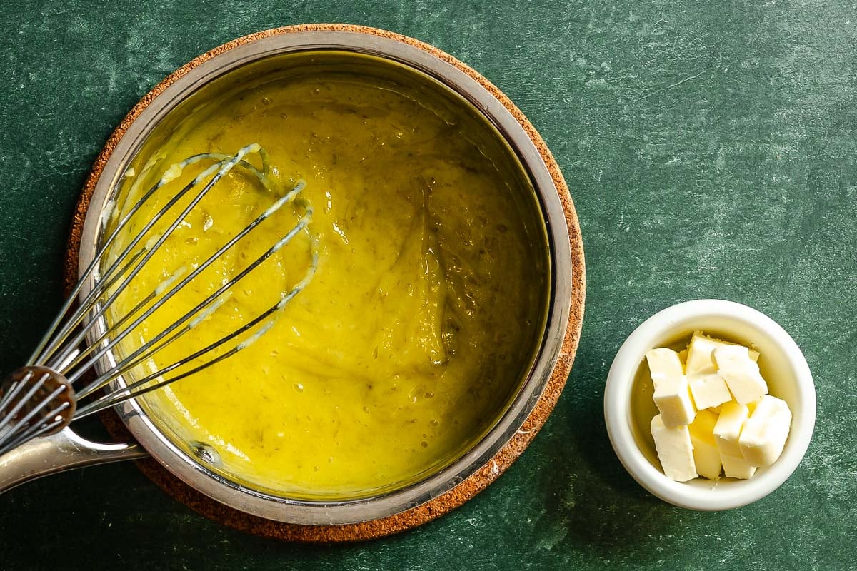 Cooked lime curd in a saucepan with a whisk set inside, butter set in a bowl nearby.