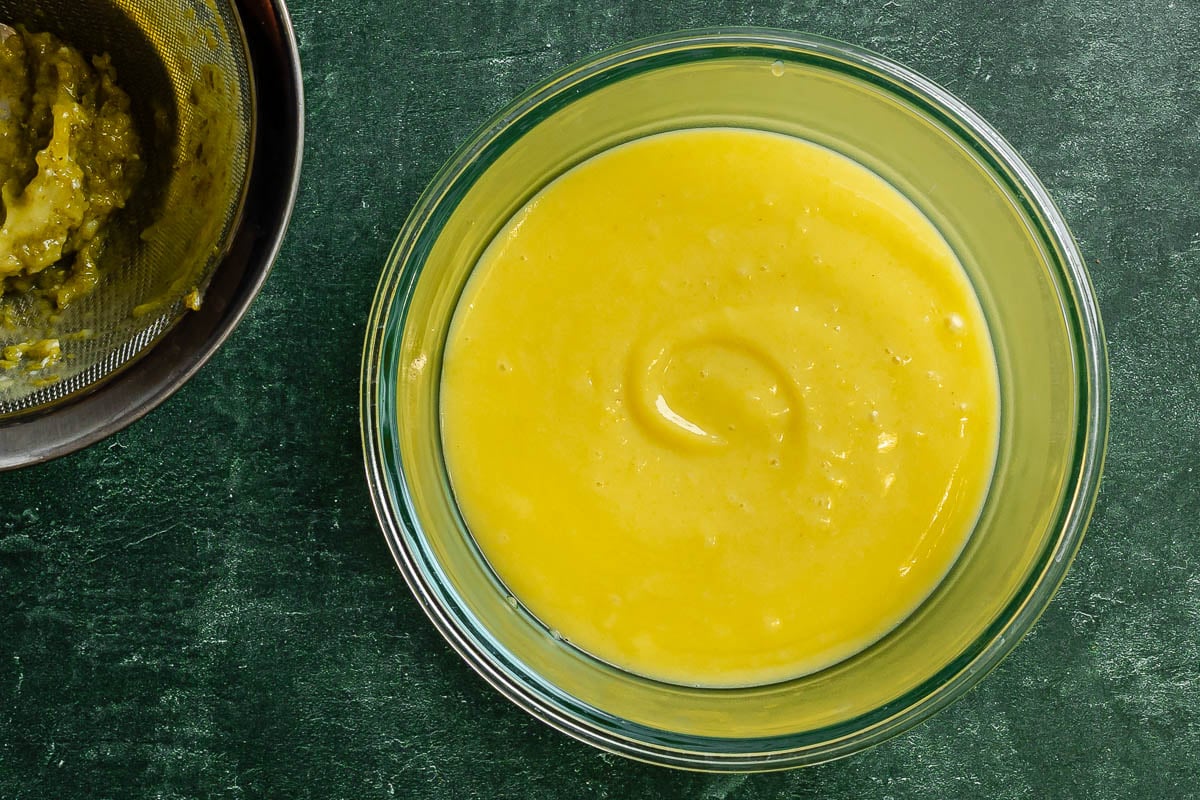 Lime curd in a glass bowl with a fine mesh sieve of strained out lime zest set nearby.