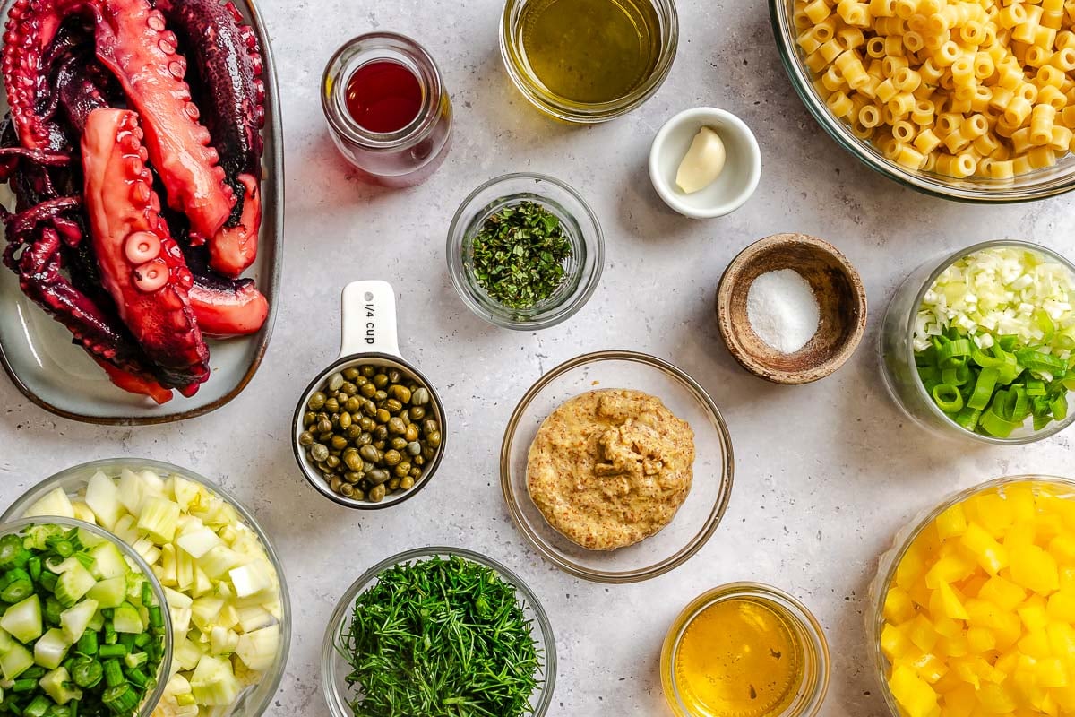 Ingredients for an octopus pasta salad arranged on a table, including octopus, pasta, capers, veggies, mustard, oil and vinegar.
