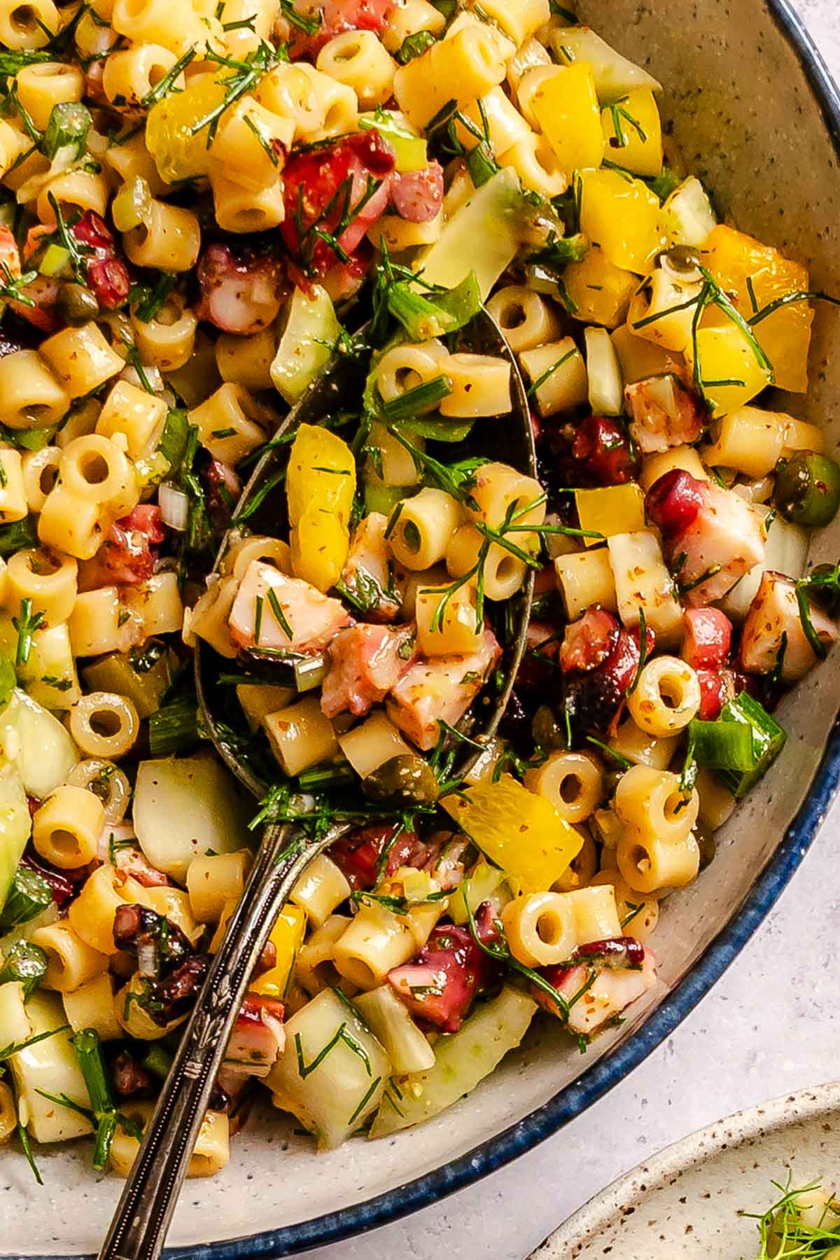 A bowl of pasta salad with diced vegetables, diced octopus, herbs, and dressing, served with a fork on a textured surface.