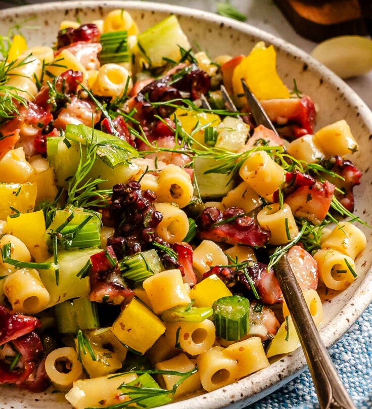 A bowl of pasta salad with diced vegetables, diced octopus, herbs, and dressing, served with a fork on a textured surface.