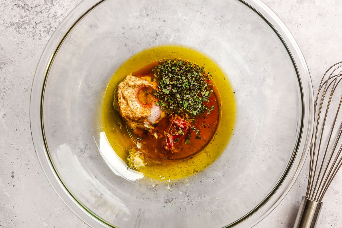 Olive oil, mustard, vinegar, and minced oregano in a large glass bowl ready to be whisked together.