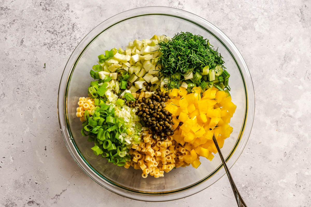 Cooked pasta, diced bell pepper, diced fennel, fennel fronds, sliced scallions and capers in a large glass mixing bowl set on a gray surface.