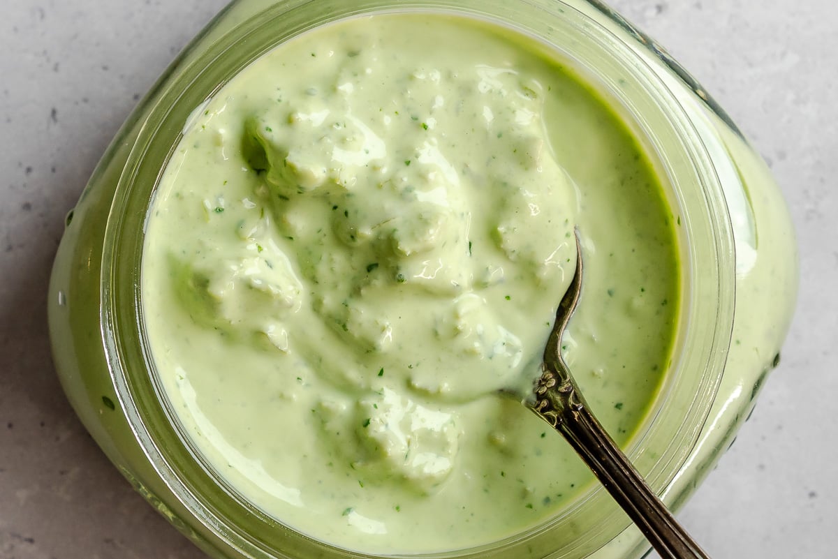 A jar of creamy green salad dressing with a spoon in it, seen from above on a light surface.