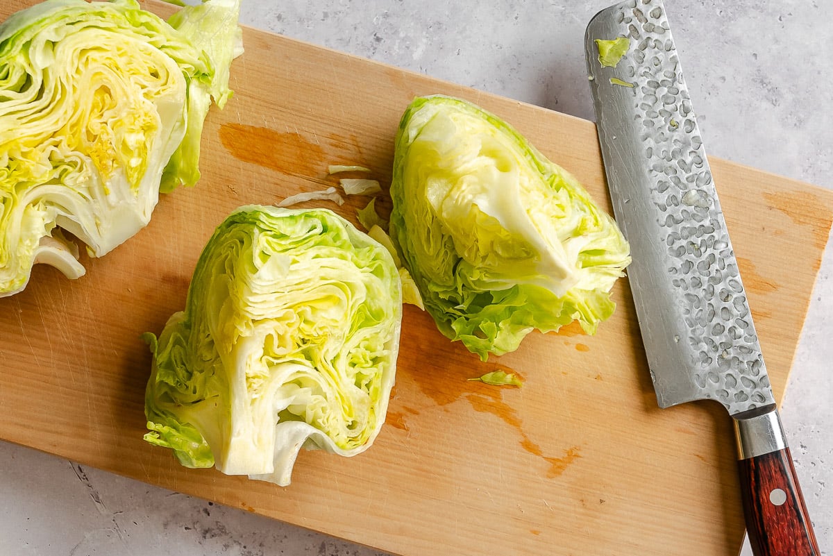 Iceberg lettuce cut in half on a wooden cutting board with a textured knife.