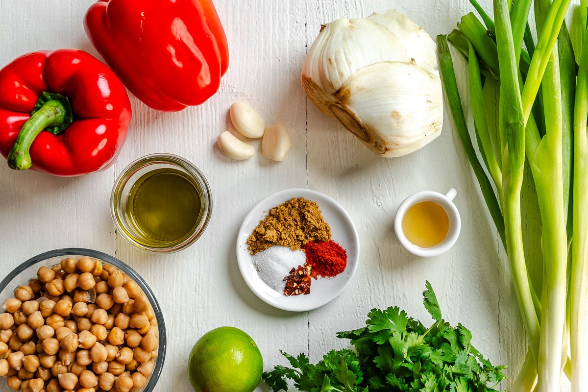 Chickpeas, red pepper, garlic, fennel, green onions, oil, spices, cilantro, and a lime on a white surface.