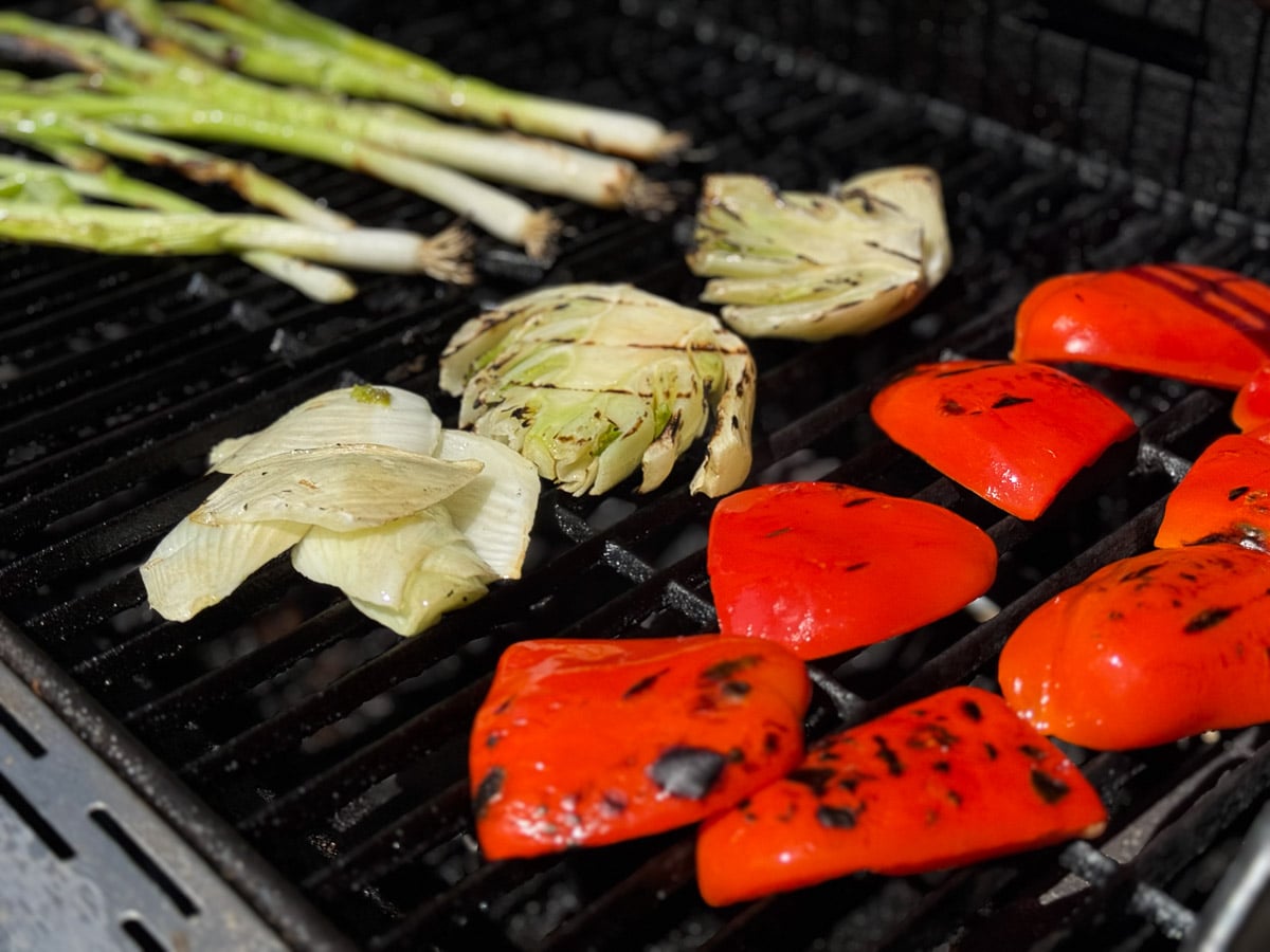 Large chunks of bell peppers, slices of fennel, and green onions charring on a grill.