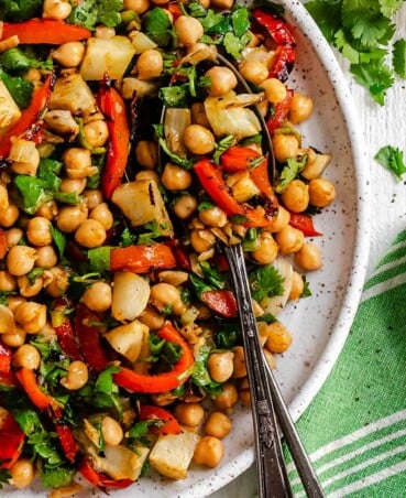 A plate of chickpea salad with grilled red peppers, fennel, cilantro, and two spoons, with lime wedges in the background.