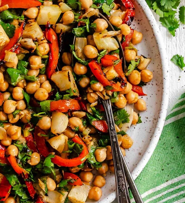 A plate of chickpea salad with grilled red peppers, fennel, cilantro, and two spoons, with lime wedges in the background.