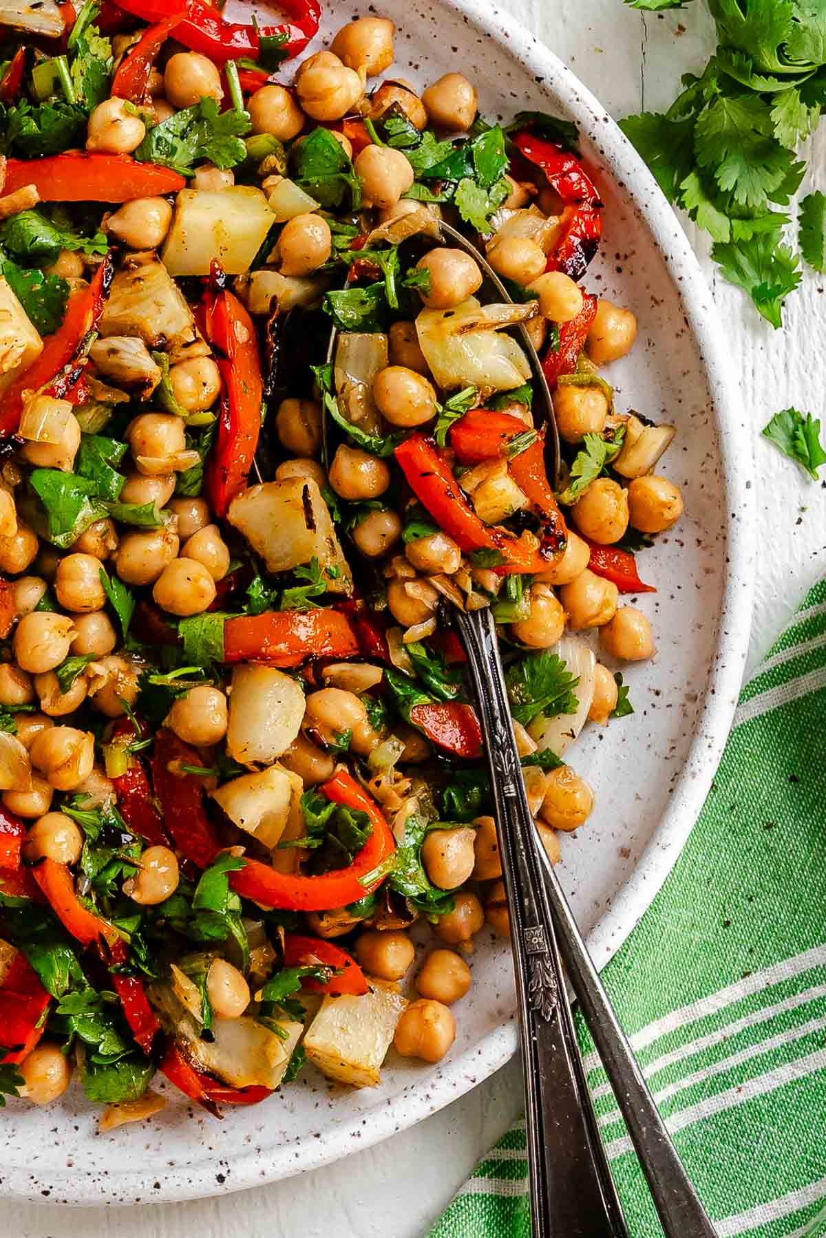 A plate of chickpea salad with grilled red peppers, fennel, cilantro, and two spoons, with lime wedges in the background.