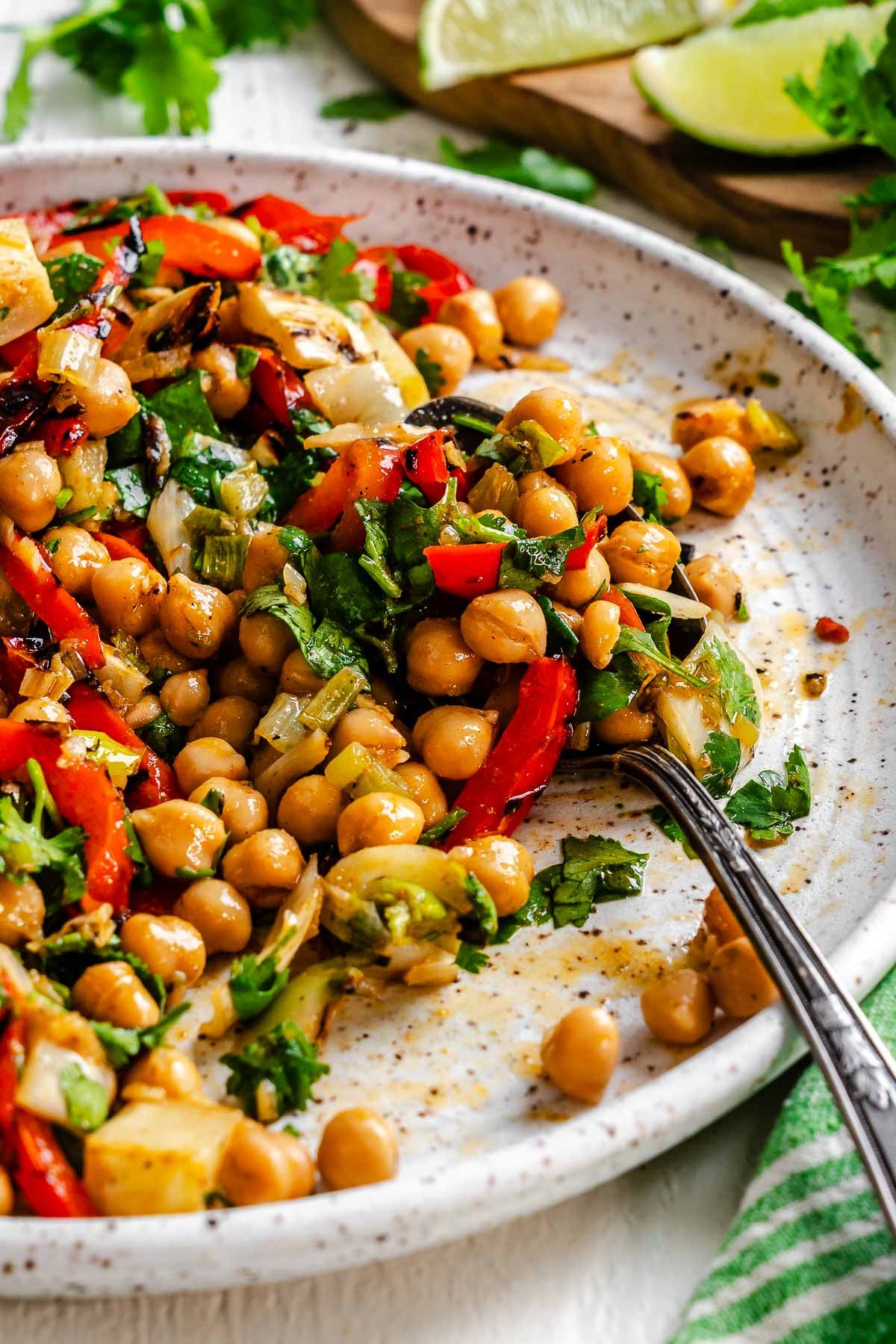 A plate of chickpea salad with grilled red peppers, fennel, cilantro, and two spoons, with lime wedges in the background.