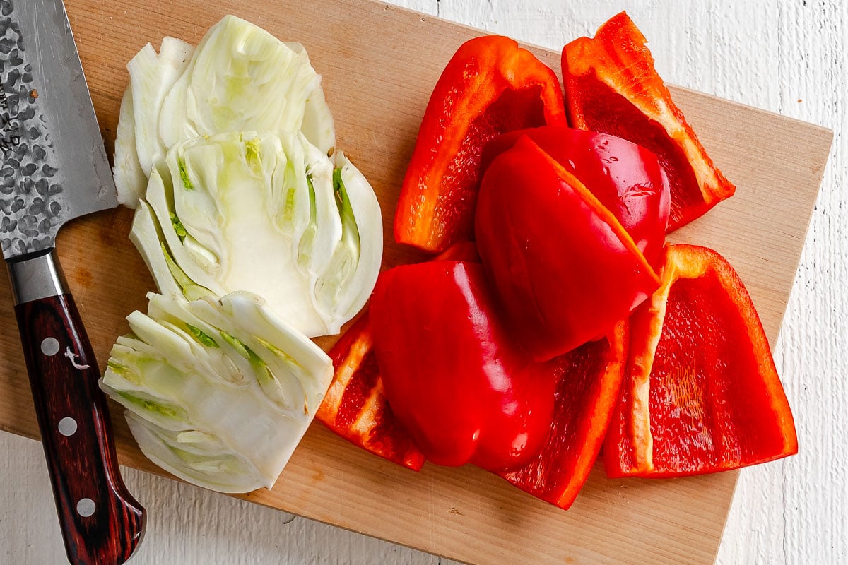 Sliced raw fennel, large chunks of red bell peppers on a wood cutting board.