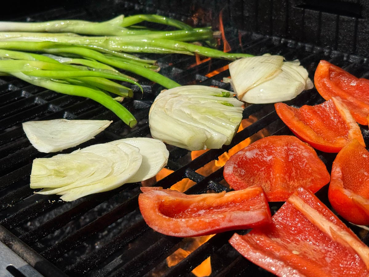 Large chunks of bell peppers, slices of fennel, and green onions charring on a grill.
