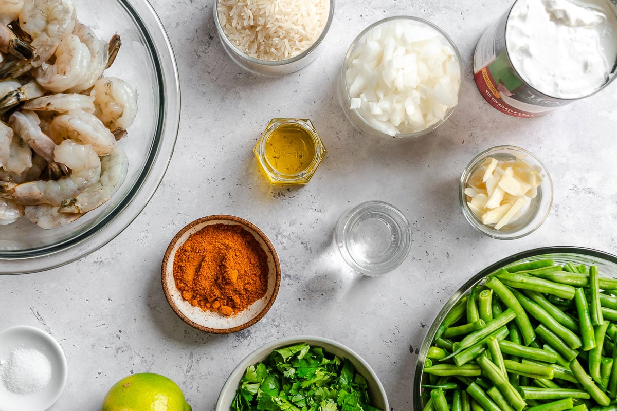 Bowls of shrimp, rice, onions, green beans, curry powder, cilantro, sliced garlic, oil, and salt set on a counter with a can of coconut milk and a lime.
