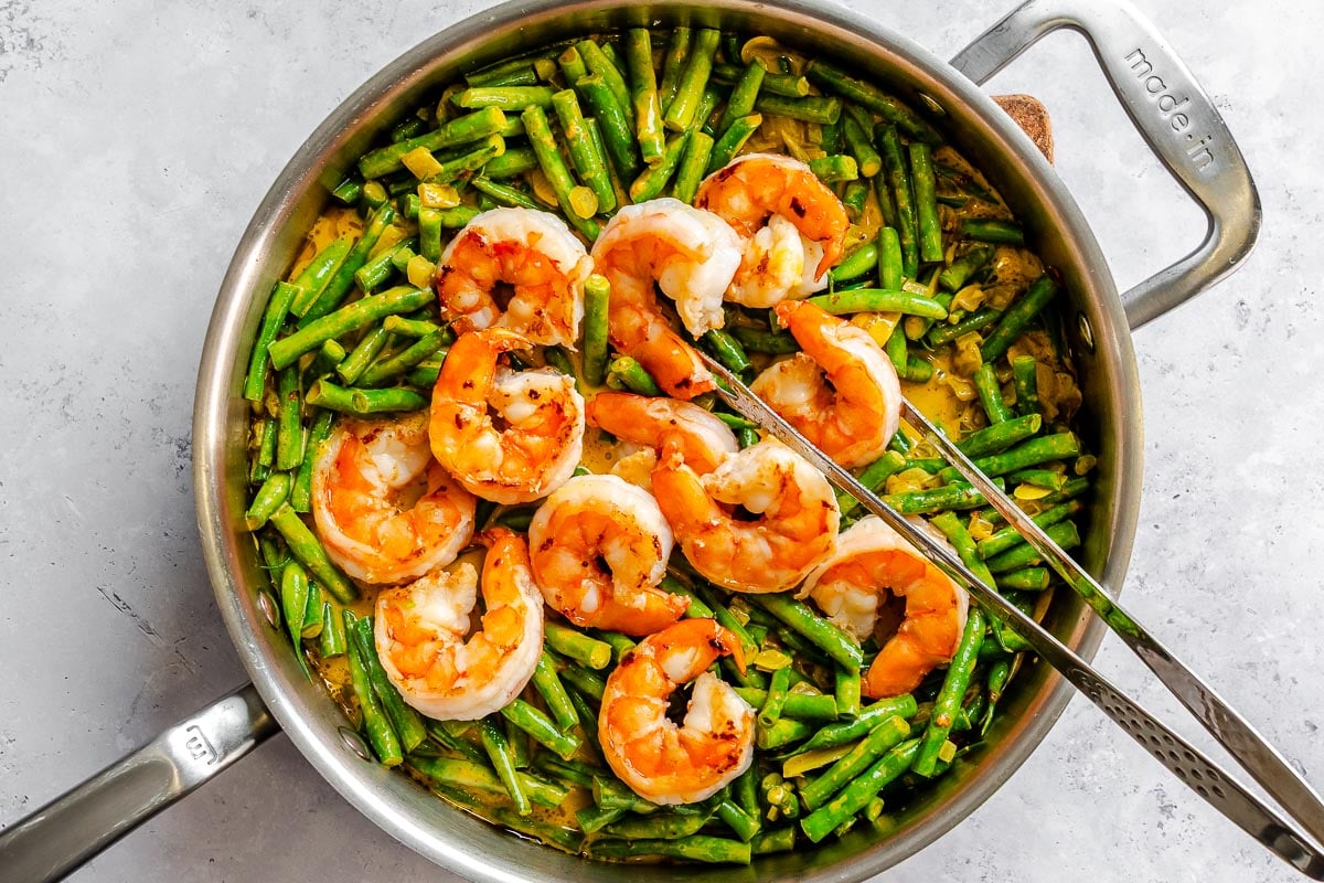 Shrimp and green beans being cooked in a stainless steel pan with curry sauce, with metal tongs resting on top.