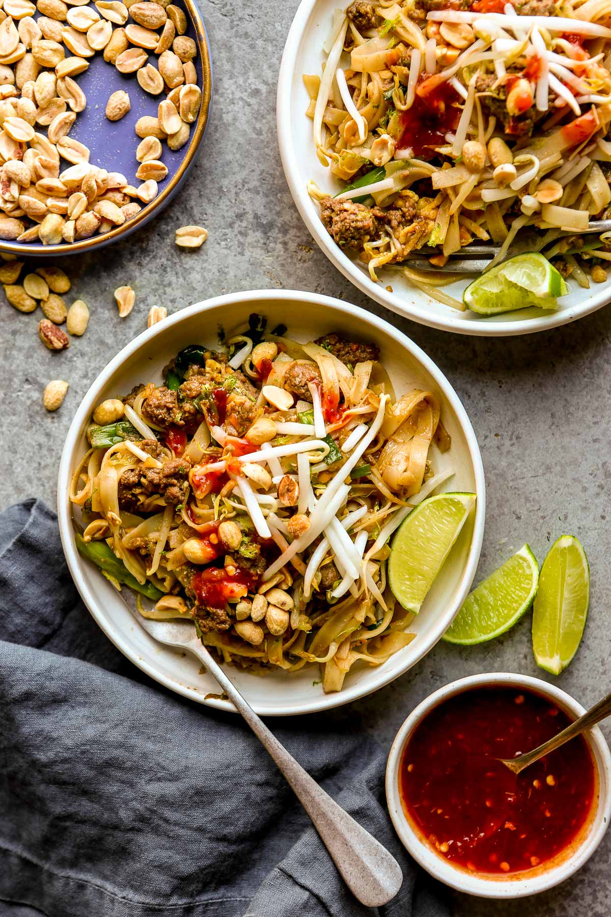 Two plates of beef pad thai with lime wedges, peanuts, and bean sprouts, plus a bowl of chili sauce on the side.